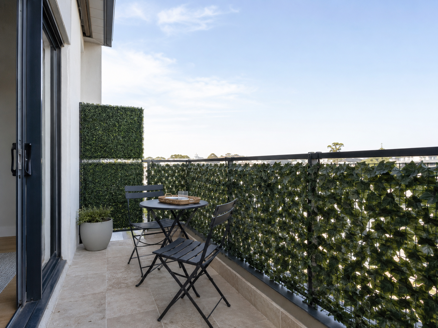 Compact balcony with artificial ivy along the railing, two square boxwood privacy panels and a black bistro set.