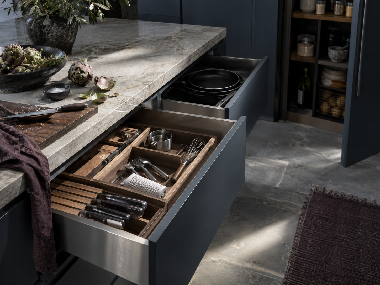 Open kitchen drawers with cooking tools and cookware positioned beside a nearby pantry cabinet.